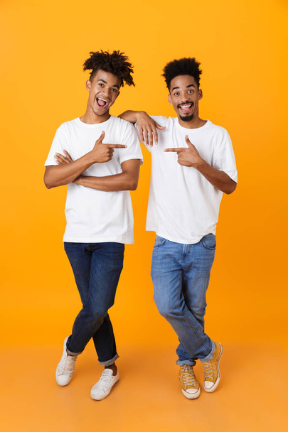 Full length of two happy male african friends in t-shirts standing isolated over yellow background, pointing fingers at each other - Foto, Imagem