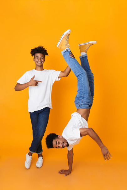 Full length of two happy male african friends in t-shirts isolated over yellow background, having fun, standing upside down - Foto, immagini