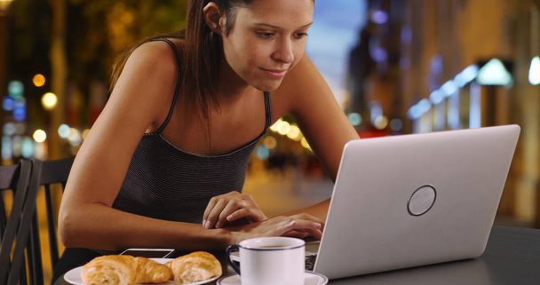 Young woman in Paris sitting outside using laptop and eating croissant - Photo, Image
