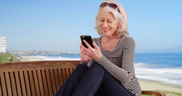 Stare biały, kobieta siedzi na ławce przez brzeg wysyłanie tekstu w telefonie - Zdjęcie, obraz
