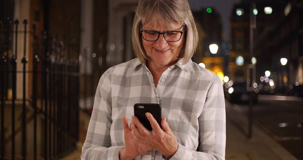Mujer mayor sonriente usando el teléfono para enviar mensajes de texto en la acera de la ciudad por la noche
 - Foto, Imagen