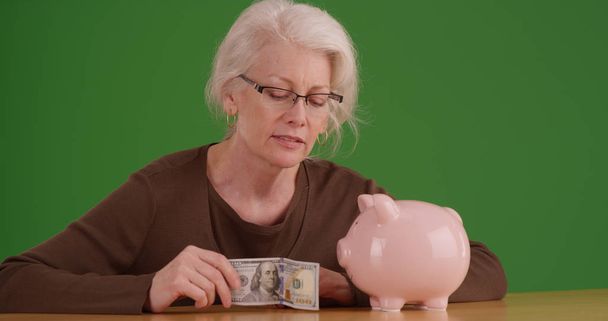 Пожилая женщина собирается положить 100-долларовую банкноту в копилку на зеленый экран
 - Фото, изображение