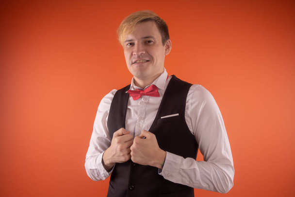 A handsome young man in a white shirt with a black vest and a red butterfly posing on an orange background - Valokuva, kuva