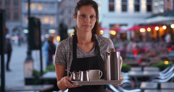 Ontevreden serveerster schort dragen en uitvoering dienblad met koffie op het terras - Foto, afbeelding