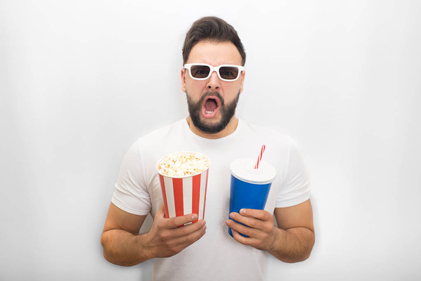 Excited man keeps his mouth opened. He holds bucket of popcorn and glass of cola at the same time. Guy wears movie glasses. Isolated on white background. - Foto, Bild