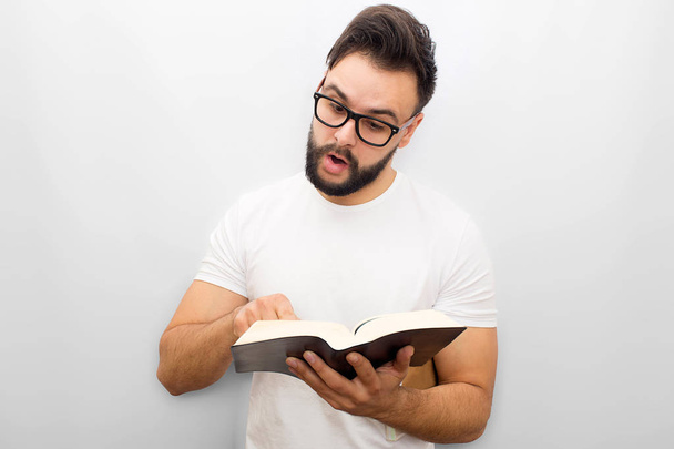 Picture of young man reading aloud from book. He holds it in hands and points on it. Young man is concentrated. Isolated on white background. - Фото, зображення