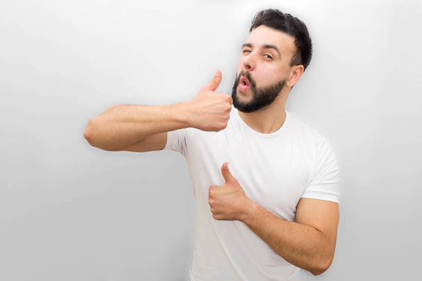 Nice and positive young man stand and winkle on camera. He shows his bog thumbs up. Guy is happy. Isolated on white background. - Φωτογραφία, εικόνα