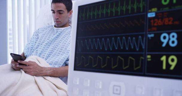 Nahaufnahme junger männlicher Latino-Patienten auf dem Handy neben dem ekg-Monitor - Foto, Bild