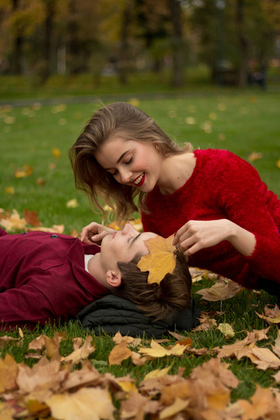 the girl and the guy in red sweaters and jeans sit in the park on the green grass in the yellow maple leaves, the girl smiles.a date in the fall.Leaves a leaf against the face of a man, protection, - Photo, Image