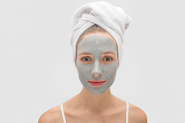 Peaceful young woman stands and looks on camera. her face is covered with mud mask. Model is calm. White towel covers her hair. Isolated on grey background. - Foto, imagen