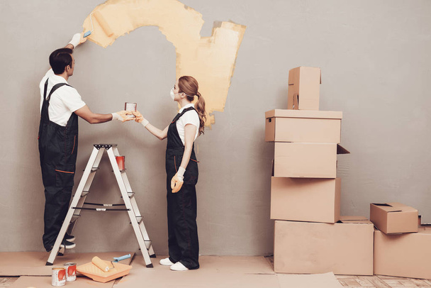 Master Painter. Gray Wall. Cardboard Boxes and Construction Ladder. Man and Woman in Uniform. Master Painter with Paint and Brush in Hand. Painting Walls. Repair Home. Gray Background. Master Painter. - Foto, Bild