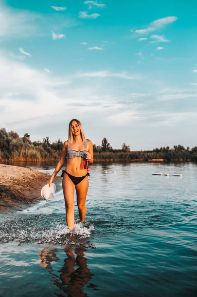 Retrato de una joven mujer impresionante en traje de baño caminando en el lago. Hermosa chica sonriente divirtiéndose en el agua mientras sostiene una botella de cerveza y un sombrero
 - Foto, imagen