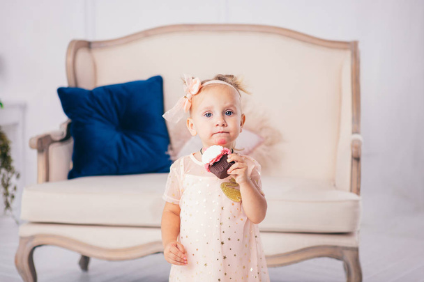 子供の誕生日。面白い2歳の白人の女の子は、家の寝室に立って、ケーキのデザートを保持し、食べて、ピンクのドレスを着ています。顔と口に塗りつぶされ、クリームで塗りつぶされた - 写真・画像