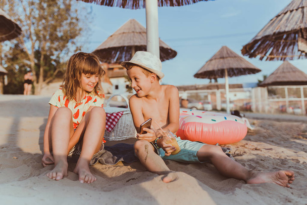 Ritratto di bambini positivi che giocano insieme con i telefoni cellulari sulla spiaggia sabbiosa. Utilizzando le applicazioni e il browser sullo smartphone
. - Foto, immagini