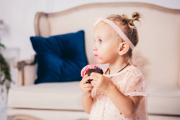 子供の誕生日。面白い2歳の白人の女の子は、家の寝室に立って、ケーキのデザートを保持し、食べて、ピンクのドレスを着ています。顔と口に塗りつぶされ、クリームで塗りつぶされた - 写真・画像