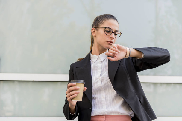 Portrait of businesswoman in front of building looking on wristwatch and drinks coffee - Photo, Image