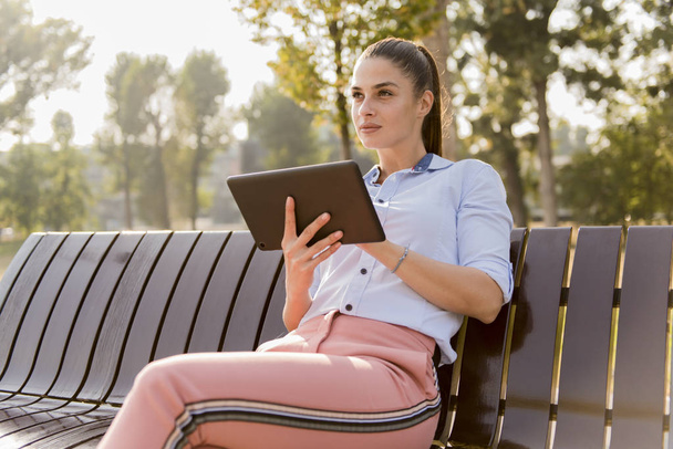 Menina bonita nova usando tablet enquanto sentado no banco do parque
 - Foto, Imagem