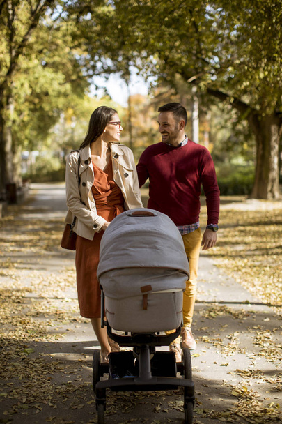 Padres jóvenes felices caminando en el parque de otoño y conduciendo a un bebé en carro de bebé
 - Foto, imagen
