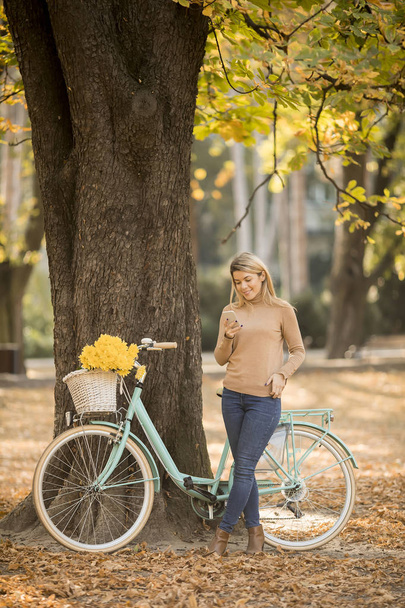 Cheerfull young woman with bicycle using smartphone in autumn park - Φωτογραφία, εικόνα