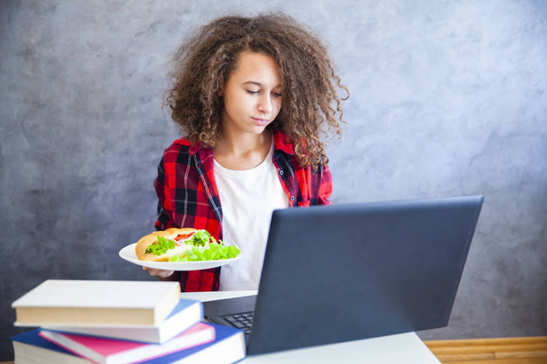 Porträt eines Mädchens mit lockigem Haar, das am Laptop arbeitet und Sandwich isst - Foto, Bild