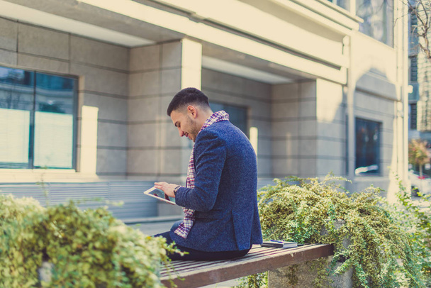 A man is sitting in front of some buildings, looking satisfied and happy at his tablet - Foto, immagini
