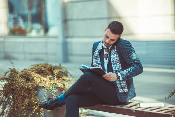 A stylish man is sitting with crossed legs on the bench, talking on his phone and writing in the notebook - Foto, afbeelding