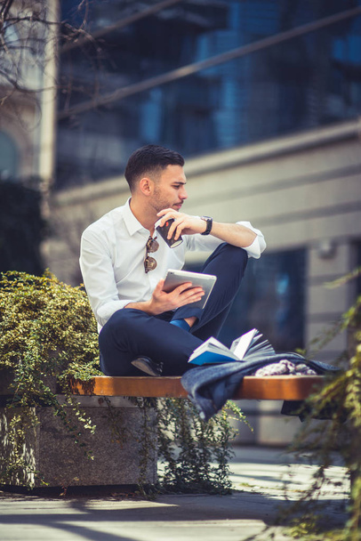 Городской бизнесмен сидит на скамейке в центре города, смотрит на свой планшет и наслаждается хорошей погодой
 - Фото, изображение