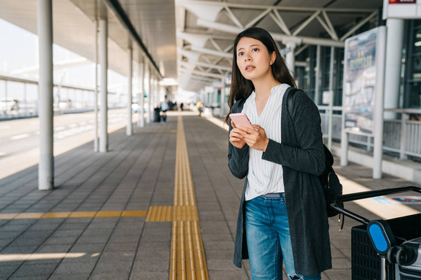 asian passenger arrive the kansai airport standing outdoor beside luggage cart on a sunny day. young girl checking the timetable of the bus to transport to kyoto. traveler self guide trip in japan. - 写真・画像