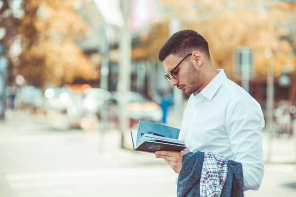 A modern young businessman in white shirt is standing in the city and looking serious while reading a book - Photo, image