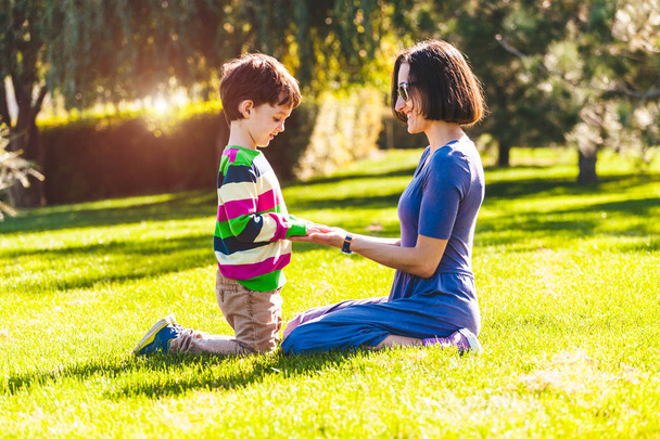 Chłopiec z jego matka siedzi na zielonej trawie. W parku chodzi dziecko z rodzicem. Matka przytula. Kobieta jest zabawy z synem w przyrodzie. - Zdjęcie, obraz