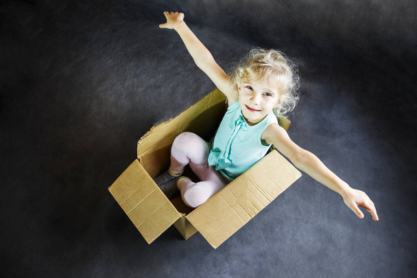 The little blonde smiles inside a cardboard box. dark background - Photo, Image