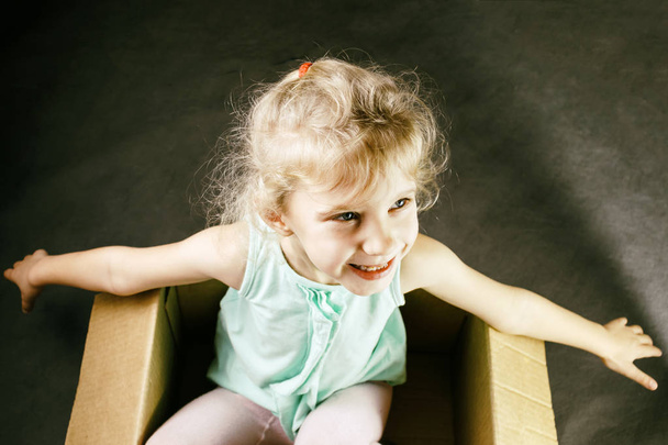 The little blonde smiles inside a cardboard box. shows us hands. dark background. toning. - Foto, imagen