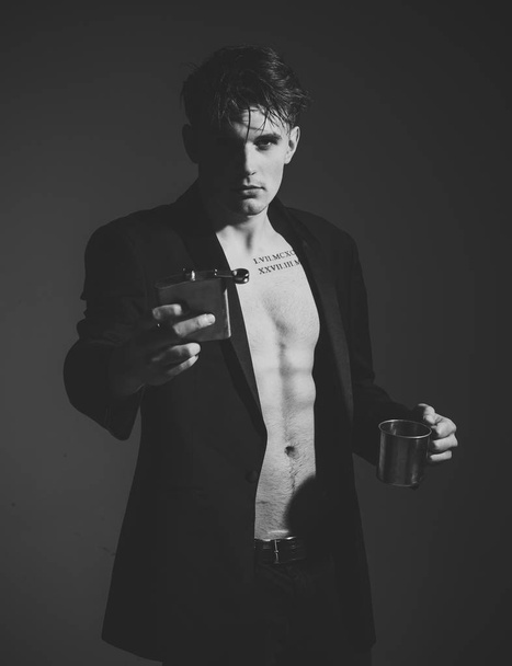 Macho on confident face, muscular figure, holds flask with alcohol and metallic cup. Luxury alcohol concept. Man with muscular torso, six packs, wears classic jacket, looks attractive, dark background - Photo, image