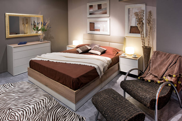Концепція дизайну двомісної спальні з високим коричневим ліжком і зручним кріслом з пуфманом, килимом зебри на підлозі і картинами на стінах
 - Фото, зображення