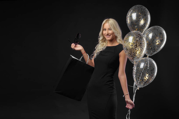 Elegant blonde woman wears sunglasses and black dress holding black shopping bags and festive balloons, black friday concept - Fotó, kép