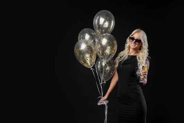 Brightfull expressions of happy emotions of amazing blonde girl celebrating party on black background. Luxury black dresses, smiling, a glass of champagne, golden tinsels, balloons, long curly hair - Foto, afbeelding