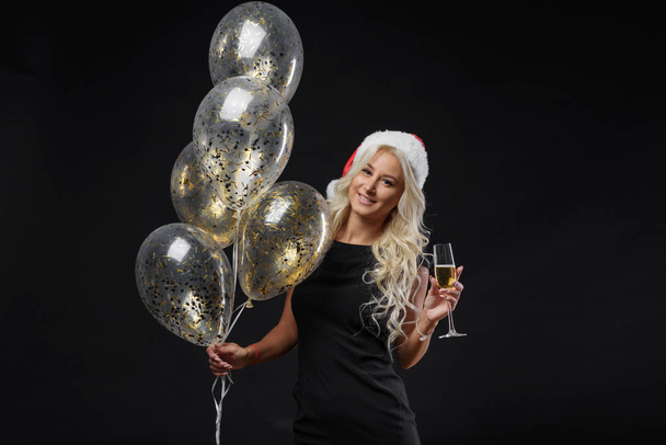Brightfull expressions portrait of happy emotions girl in Santa hat celebrating Christmas party on black background. Holds a glass of champagne and golden balloons. New year holiday concept - Foto, Bild