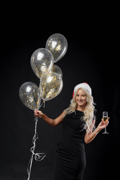Brightfull expressions portrait of happy emotions girl in Santa hat celebrating Christmas party on black background. Holds a glass of champagne and golden balloons. New year holiday concept - 写真・画像