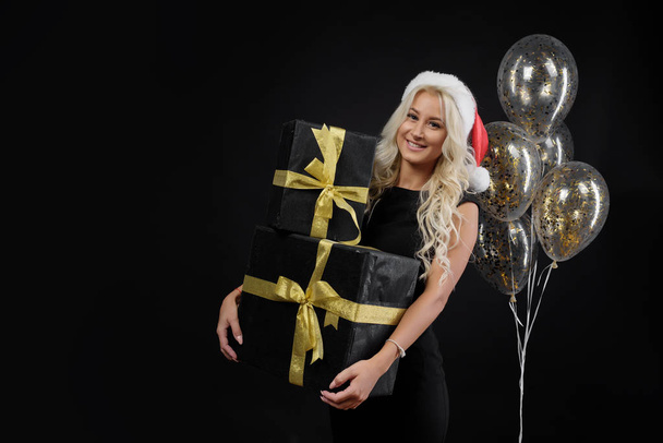 Portrait of beautiful smiling woman in black dress and Santa hat holding stack of gift boxes on the black background. Holding golden balloons and Celebrated a Christmas holiday. Sale concept - Foto, Imagem