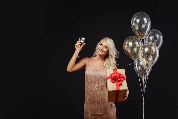 Joyful young woman in luxury dresses celebrating birthday or Christmas party with confetti on black background.Having fun and smiling. Holding present, gift, golden balloons and glass of champagne - Foto, afbeelding