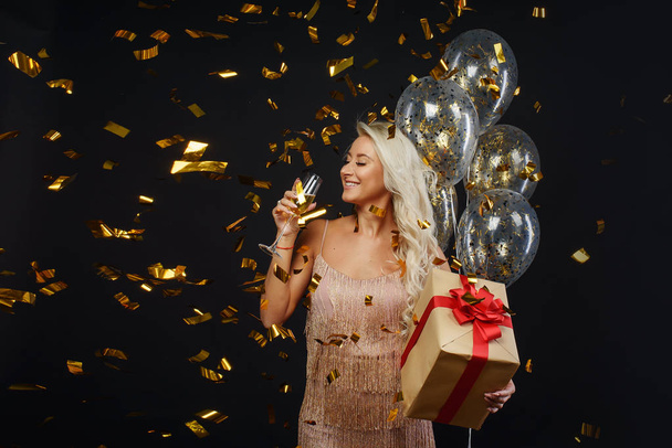 Joyful young woman in luxury dresses celebrating birthday or Christmas party with confetti on black background.Having fun and smiling. Holding present, gift, golden balloons and glass of champagne - Photo, image