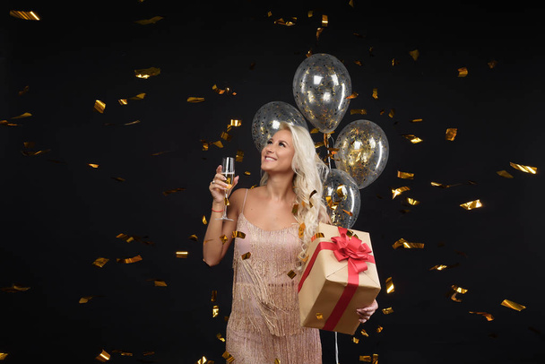 Joyful young woman in luxury dresses celebrating birthday or Christmas party with confetti on black background.Having fun and smiling. Holding present, gift, golden balloons and glass of champagne - Photo, Image