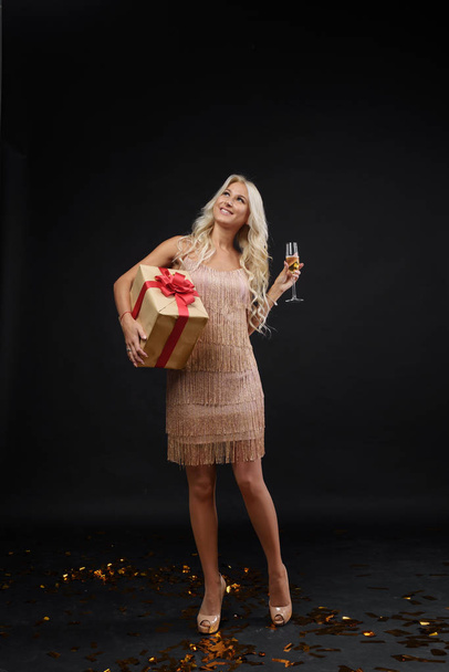 Joyful fashionable young woman in luxury dresses celebrating birthday or Christmas party on black background.Having fun and smiling. Holding present, gift and glass of champagne - 写真・画像