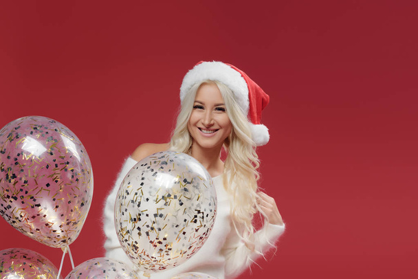 Christmas party. Happy excited young woman in santa claus hat with balloons over red background - 写真・画像