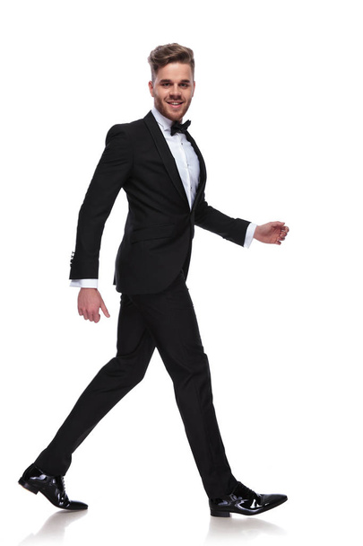 laughing young man in tuxedo and bow tie walks and looks at the camera on white background - Φωτογραφία, εικόνα