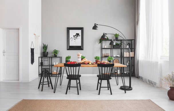 Black chairs at dining table with food in apartment interior with lamp and poster on grey wall. Real photo - 写真・画像