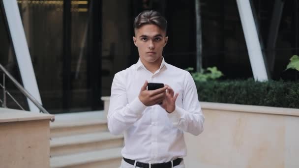 Portrait of young successful Businessman with smartphone standing near office building and looking into the camera. Concept: new business, travel the world, communication, contacts, business deal - Séquence, vidéo