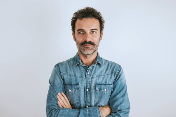 Portrait of brown, smiling, handsome man with mustache in jeans shirt standing with crossed arms. Gray background. - Foto, Imagen