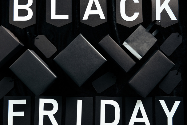 top view of black friday lettering, blank price tags, shopping bag and boxes on black background - Фото, изображение