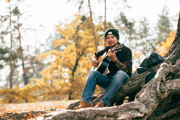 dorosły mężczyzna gra na gitarze akustycznej, siedzi na drzewie w parku  - Zdjęcie, obraz